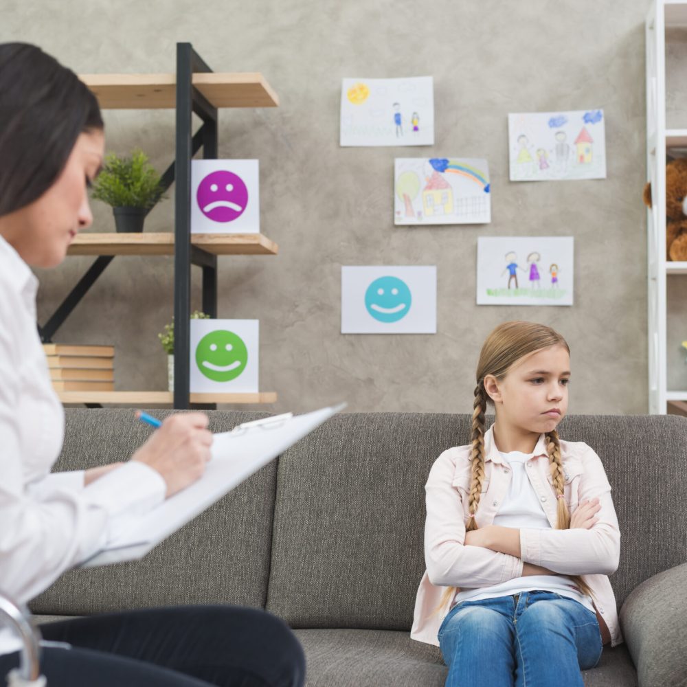 depressed-girl-sitting-sofa-with-female-psychologist-writing-note-clipboard