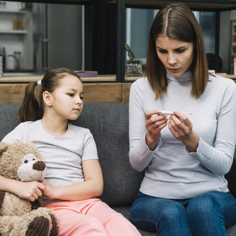 girl-sitting-gray-sofa-holding-teddy-bear-looking-woman-checking-temperature-thermometer